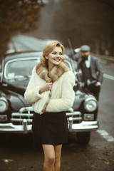 sexy woman in fur coat and bearded man. Retro collection car and auto repair by mechanic driver. Travel and business trip or hitch hiking. Couple in love on romantic date. Escort of girl by security.