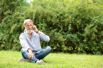 Handsome mature man with mobile phone sitting on green grass in park
