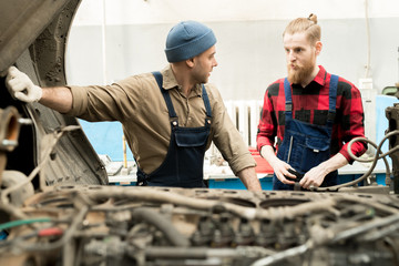 Young male car mechanic standing by broken truck and consulting with experienced bearded colleague about repair