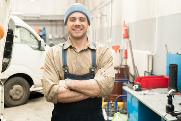 Portrait of young Caucasian male auto mechanic standing in repair shop with crossed hands and smiling at camera cheerfully