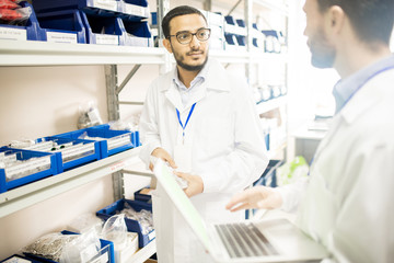 Serious confident young Arabian factory lab technician in eyeglasses wearing badge keeping records of fasteners in warehouse while discussing assemble of new device with colleague
