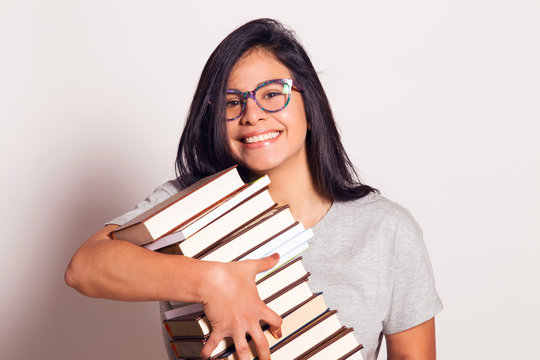 Latin nerdy girl holding in hands a pile of books. Indoors, over a white wall.