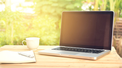 close-up laptop with cup put on a wooden slab against  natural background.  Laptop put on an outdoor table with light orange sunshine.