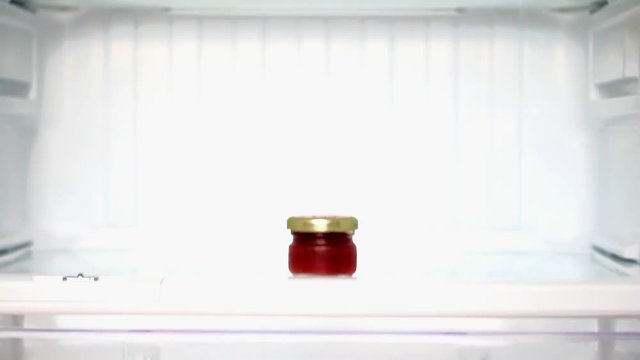 The delicates in a small glass in an empty refrgerator. A small bottle of jam in a shelf in the fridge.