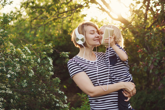 Mother and son in striped T-shirts. Woman listens to music on headphones and holds boy in her arms. He holds cell phone and kisses his mother. Family time together.