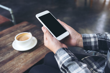Mockup image of a woman's hands holding white mobile phone with blank black desktop screen with coffee cup on wooden table in cafe