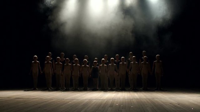 A large group of children of the dancers synchronously go on stage in the dark on a black background. Group of children engaged in ballet on stage, portrait. Slow motion. Silhouette.