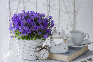 on white background purple flowers in a white basket, alarm clock, book, cup and statuette of cats