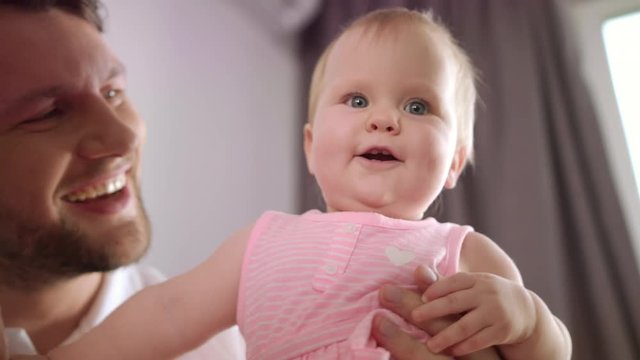 Adorable baby girl in father embrace. Closeup of cute child playing with parents. Portrait of infant playful with mom and dad. Sweet childhood. Cheerful kid with parents. Happy family with little kid