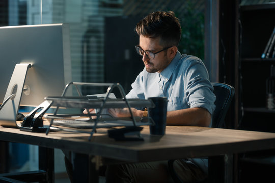 Young man in glasses working late alone at desktop in office taking notes and reading information. 