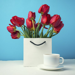Bouquet of brightly red tulips in a white gift bag, white cup with coffee on a blue background. Concept congratulations, surprises and gifts