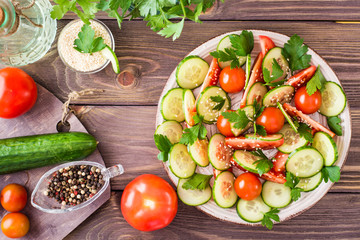 Vegetable salad of fresh cucumbers, tomatoes, parsley and sesame seeds on a plate on a wooden table. Ingredients for salad preparation. Top view