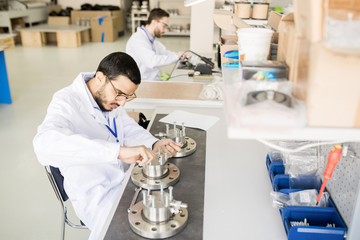Fototapeta na wymiar Concentrated busy young male engineer in lab coat and glasses focusing on bearing production and tightening bolts with hands while sitting at table