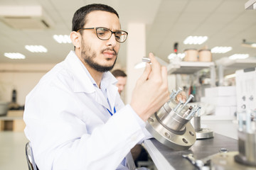 Serious frowning handsome young Arabian male engineer in lab coat viewing detail of bearing while assembling technical equipment in workshop