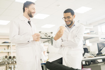 Cheerful excited young multiethnic colleagues in lab coats drinking coffee from disposable cup while talking and laughing in measuring device production shop