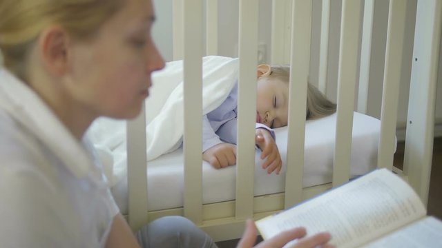 An adult woman reads a book sitting next to a sleeping little baby in a baby crib