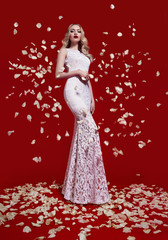 A beautiful slender girl with blond hair in a white dress stands on a red background on white rose petals.