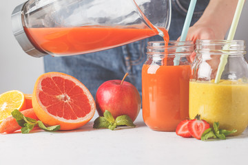 Woman cooking multicolored summer fruit juices or smoothie in glass jars and ingredients on white table.