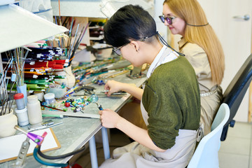 Side view portrait of two modern female artists working with glass shaping it while making beads for beautiful handmade jewelry in glassworking studio