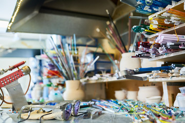 Empty workplace with various colorful glass rods on shelves by workstation in modern glassworking studio, copy space