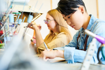 Side view portrait of young Asian female artist drawing sketches sitting at workstation with colleague busy working in creative studio, copy space