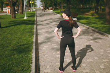 Back view Young athletic beautiful brunette girl in black uniform, cap standing, resting and looking aside before or after running, training in city park outdoors. Fitness, healthy lifestyle concept.