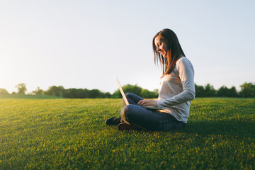 Young smart student female in casual clothes. Woman sitting on grass ground, working on laptop pc computer in city park on green grass sunshine lawn outdoors. Mobile Office. Freelance business concept