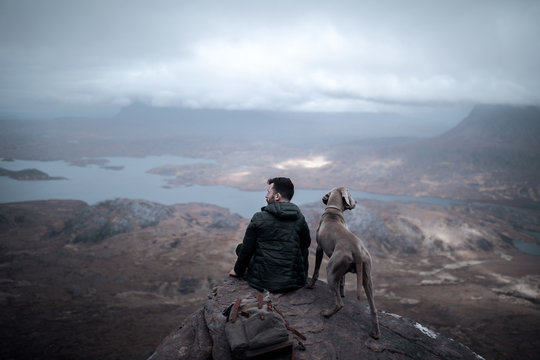 Rear view of man with Weimaraner dog sitting on rock against cloudy sky