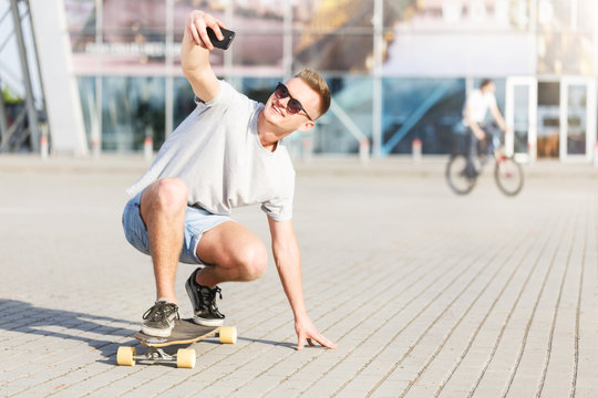 Skateboarder makes selfie on skateboard, low ride position before modern building