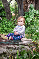 Beautiful baby sitting, smiling and posing, portrait. Little cute girl is playful in garden. Child is playing outside in park, surrounded by beautiful green trees and bushes, natural environment.