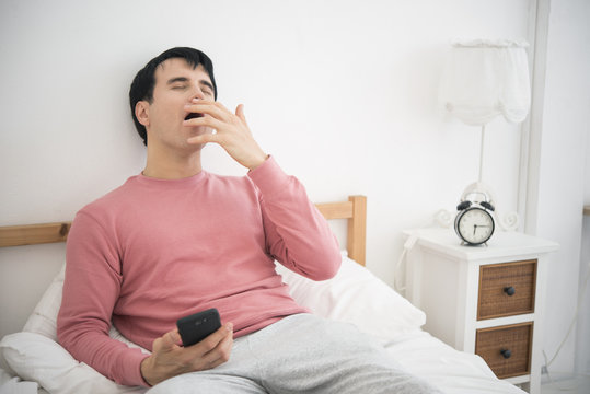 Man Playing Mobile Phone. He Sleepy On Bed In Bedroom And Alarm Clock. Sleep Concept.