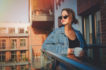 Young redhead girl in jeans clothes and cup of coffee standing on balcony alone.