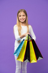Kid girl with long hair fond of shopping. Girl on smiling face carries bunches of shopping bags, isolated on white background. Girl likes to buy fashionable clothes in shopping mall. Shopping concept