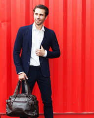 Fashion & Style. Young businessman in suit with big bag posing against red wall in sports spot.