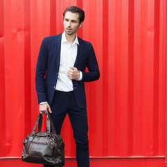 A young fashionable business man in a trendy suit posing with a large bag against the background of a red sea container.