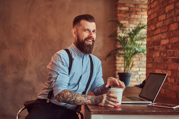 Happy handsome tattooed hipster in a shirt and suspenders holds takeaway coffee while sitting at the desk, working on a laptop in an office with a loft interior. - obrazy, fototapety, plakaty