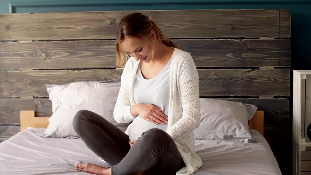 Cheerful pregnant woman massaging her belly at bedroom