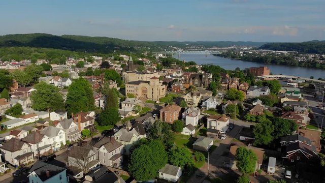 A slowly rising aerial establishing shot of a residential neighborhood in a small Pennsylvanian town. The business district and Ohio River is in the distance. Pittsburgh suburbs.  	