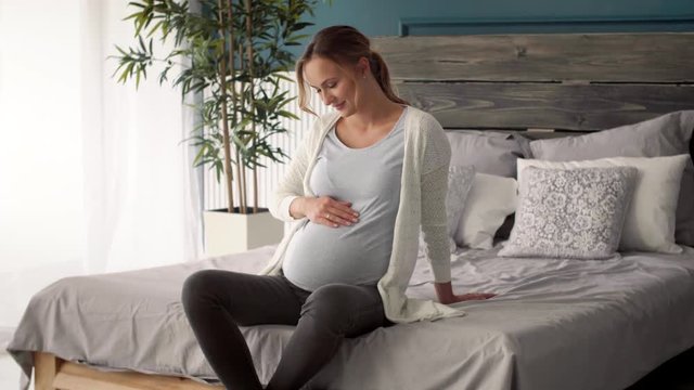 Cheerful pregnant woman touching her belly at bedroom  