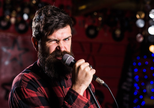 Musician with beard and mustache singing song in karaoke. Rock star concept. Man with enthusiastic face holds microphone, singing song, karaoke club background. Guy likes to sing in aggressive manner.