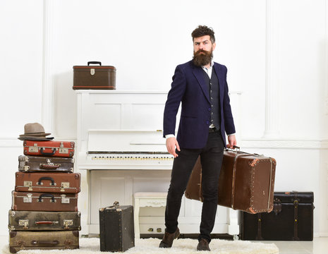 Luggage and vacation concept. Man, traveller with beard and mustache with luggage, luxury white interior background. Macho elegant on strict face stands near pile of vintage suitcase, holds suitcase.