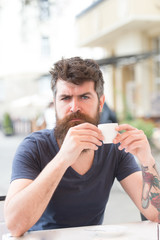 Coffee break concept. Man with long beard looks serious. Man with beard and mustache on strict or serious face, urban background, defocused. Bearded man holds espresso cup, drinks coffee at terrace
