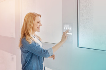 Fototapeta na wymiar Entering password. Young emotional programmer smiling and touching the transparent screen of her modern device on the wall while standing near the window