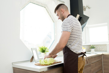 Positive busy handsome young Caucasian man with stubble wearing casual clothing and apron cutting Chinese cabbage in wooden board while preparing healthy salad in kitchen
