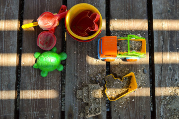 children's accessories for playing in the sand on the beach - a bucket, paddles, molds for creativity, a typewriter. They are laid out on a wooden floor in the shade. A ship made of bek