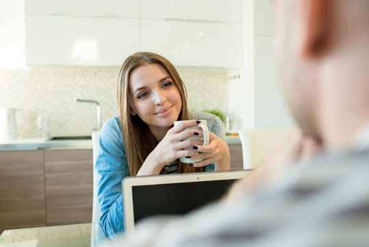 Positive Attractive Young Woman With Straight Hair Holding Mug And Attentively Listening To Husband While They Sitting In Dining Room