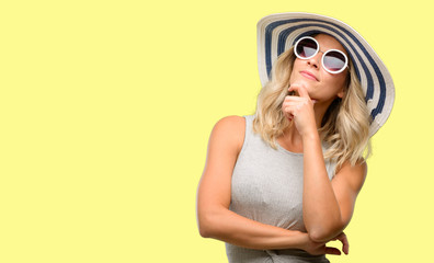 Young woman wearing sunglasses and summer hat thinking and looking up expressing doubt and wonder
