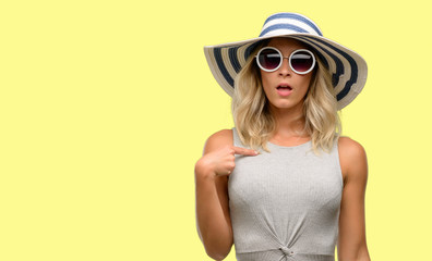Young woman wearing sunglasses and summer hat happy and surprised cheering expressing wow gesture, pointing with finger - obrazy, fototapety, plakaty
