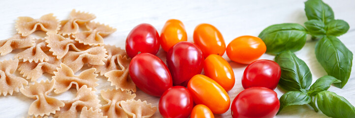Panoramic view on whole-grain farfalle, cherry tomatoes and fresh basil on a white wooden table
