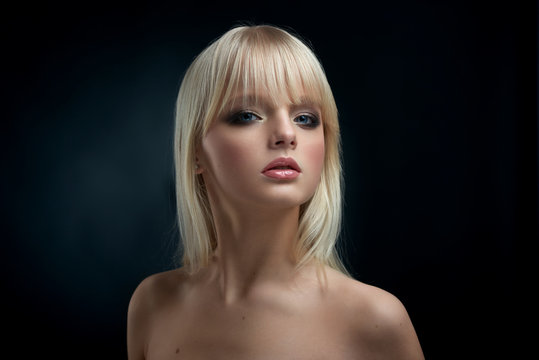 Frontview of young model with blonde hair and short haircut with opened shoulders. Girl having big blue eyes, plump lips, nice evening make up. Looking at camera, posing on black studio background.